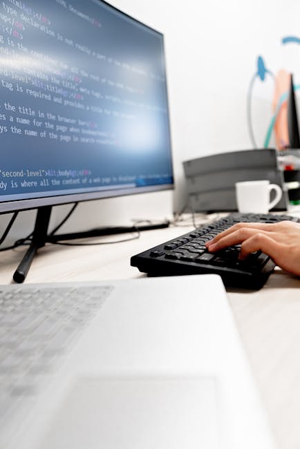 A person working on a laptop displaying lines of code, with their hands positioned over the keyboard. The laptop screen shows a dark-themed coding interface with colorful syntax highlighting. The scen