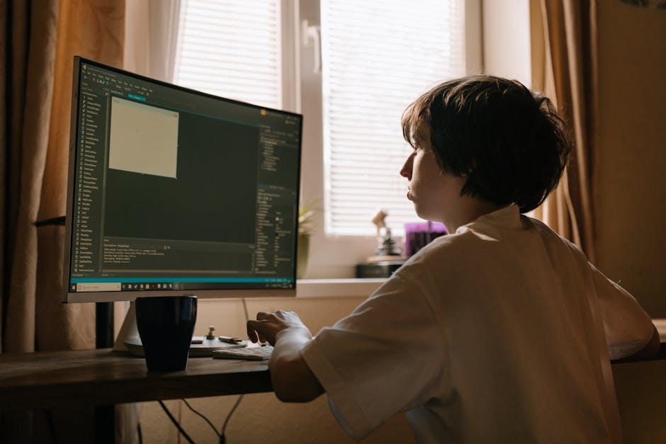 A woman with short dark hair sitting at a wooden desk in a room with natural light coming through partially open blinds. She is working on a computer with a large monitor displaying a dark-themed codi